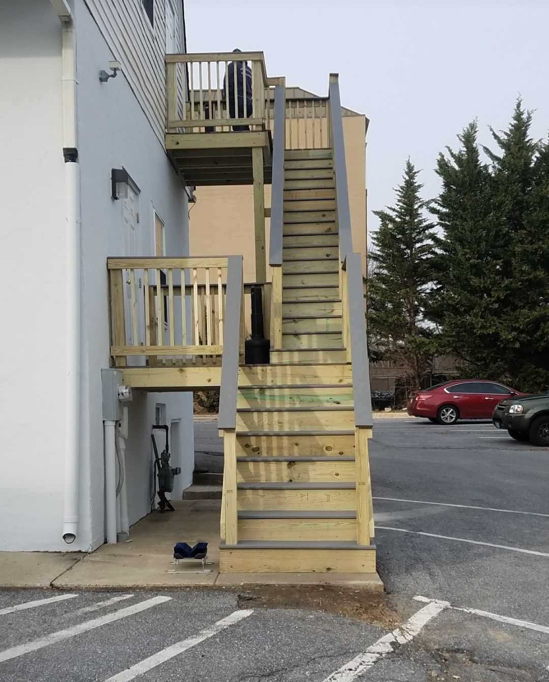 An exterior wooden staircase leads to a second-story deck on the side of a white building in a parking lot.