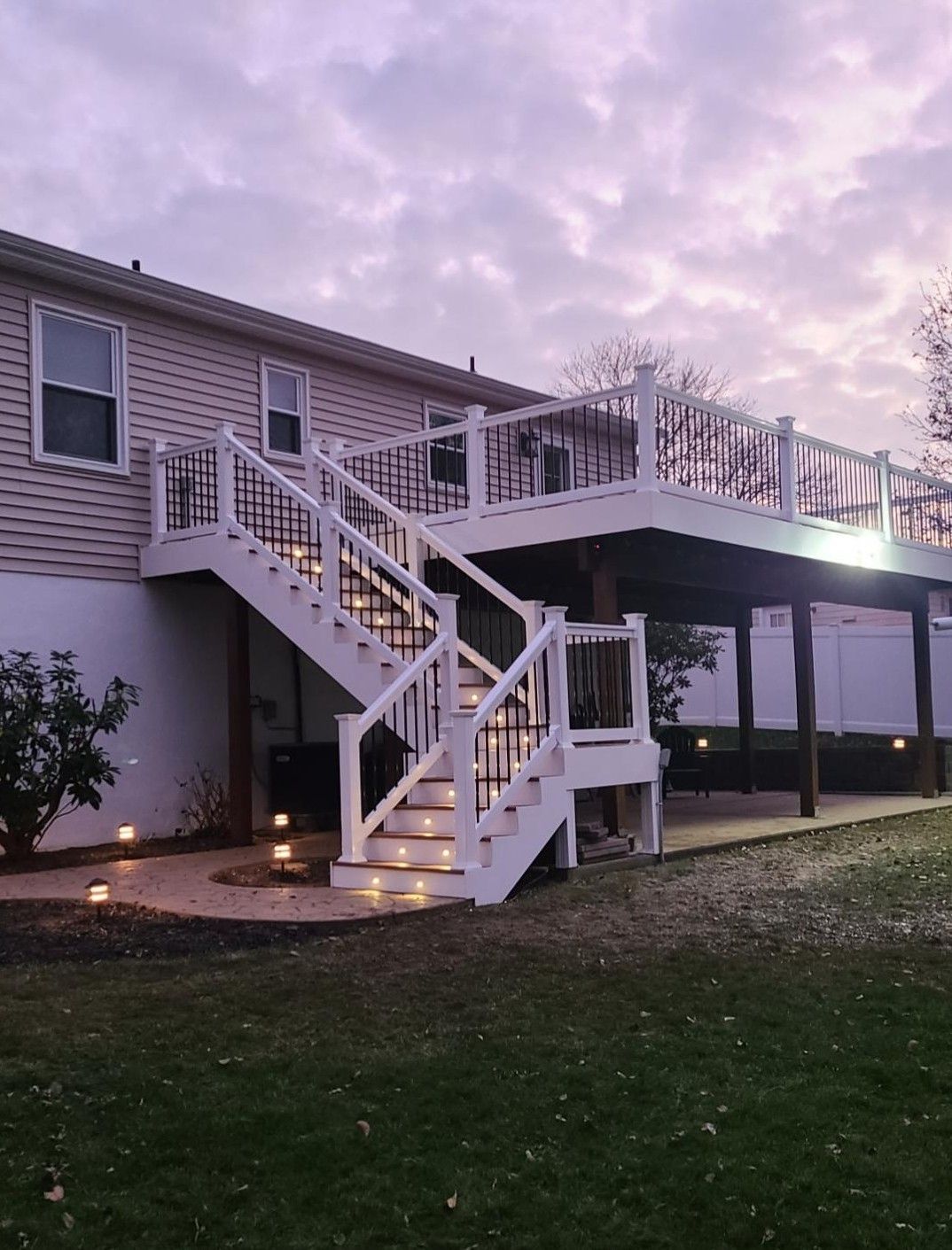An elevated back deck with white railings and illuminated stairs, featuring a patio area beneath at dusk.