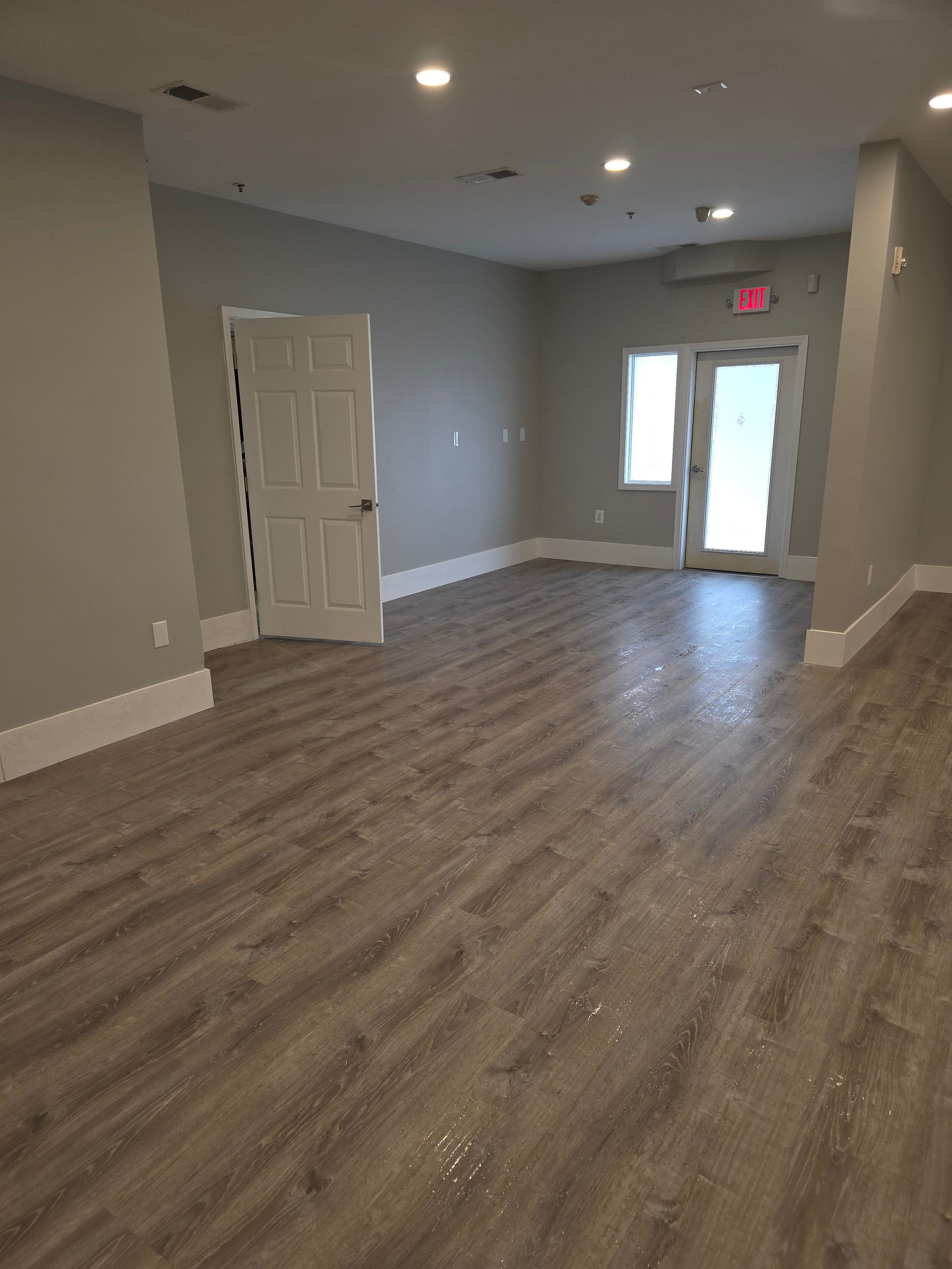A bright, empty room with grey walls, white trim, and wood-patterned flooring, featuring a white door and glass door.