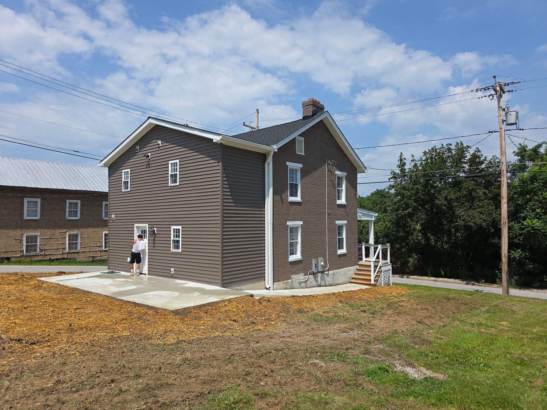 A two-story, dark brown house with white trim and a concrete patio on a sunny day with a nearby utility pole and trees.