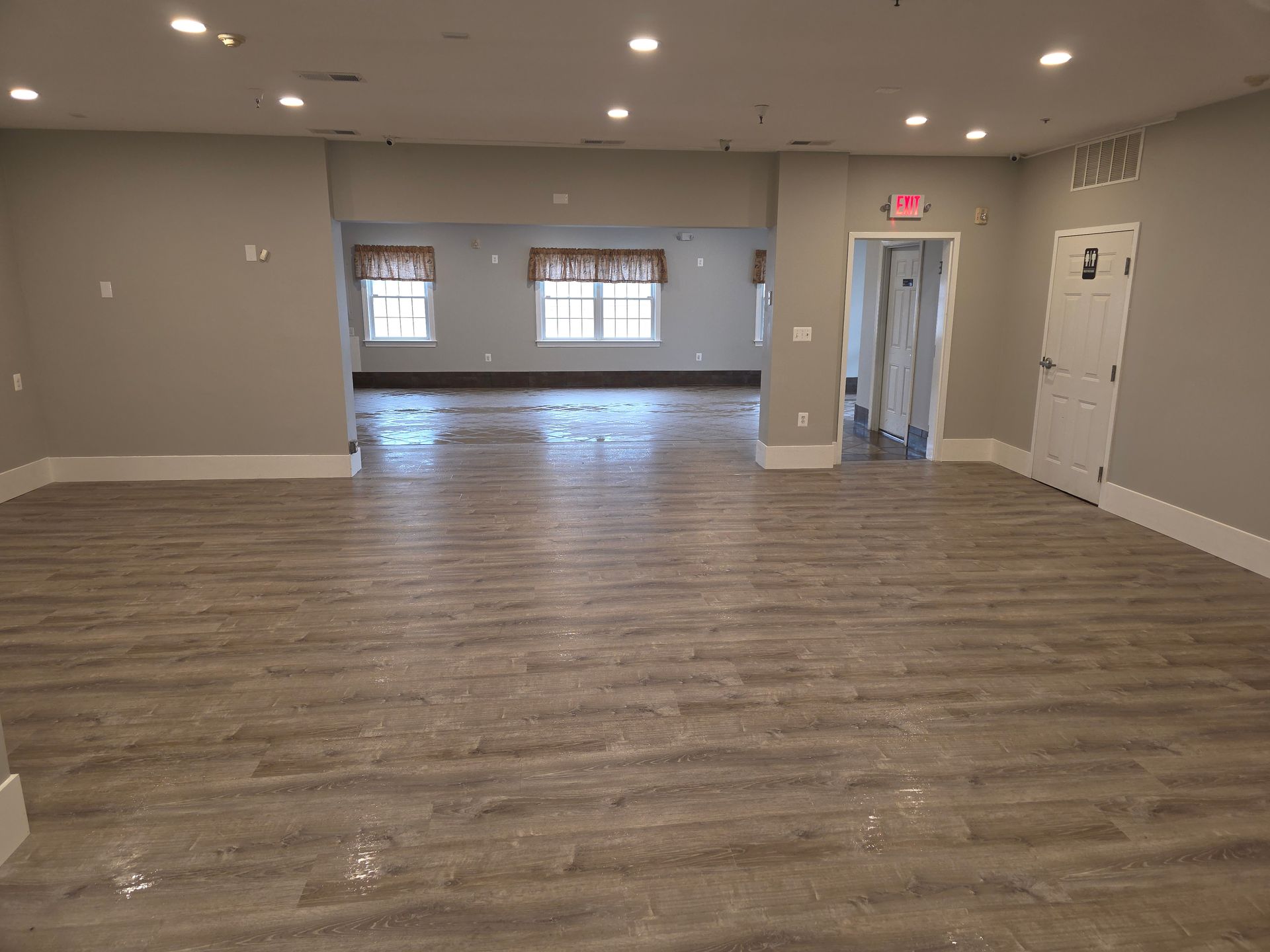 An open-plan room with grey walls, light wood-look flooring, recessed ceiling lights, two windows, and a white door.