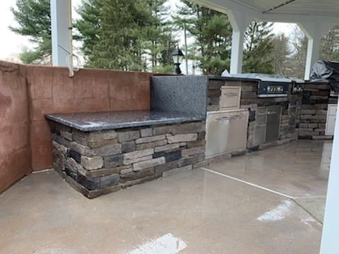 An outdoor kitchen with stacked stone siding and a dark granite countertop on a concrete patio under a covered porch.