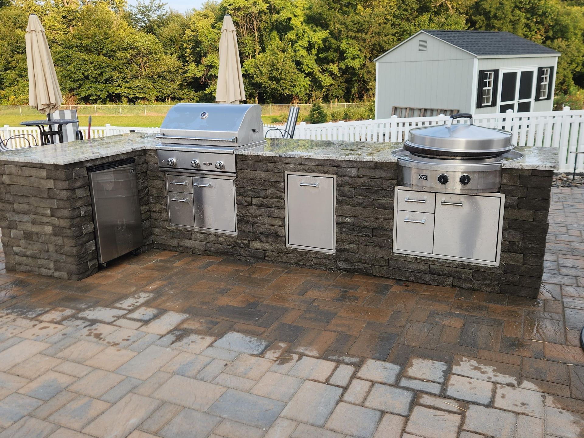 An outdoor kitchen with stainless steel appliances and stone base on a brick patio, set against a yard and white fence.