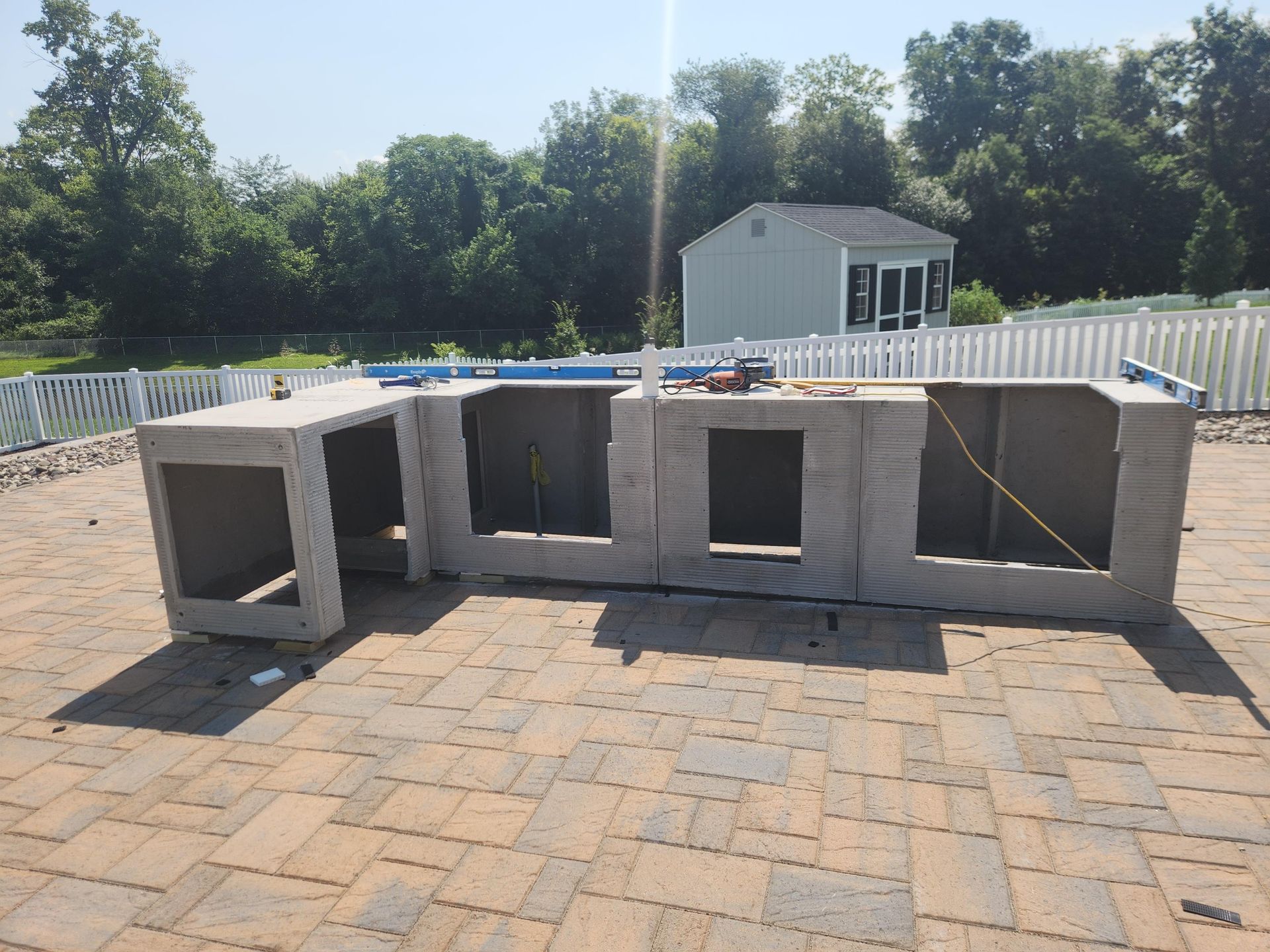 An L-shaped outdoor kitchen structure made of grey concrete blocks sits on a patio, viewed on a sunny day.