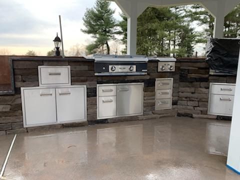 Outdoor kitchen with stainless steel appliances, drawers, and cabinets built into a stone wall under a white pavilion.