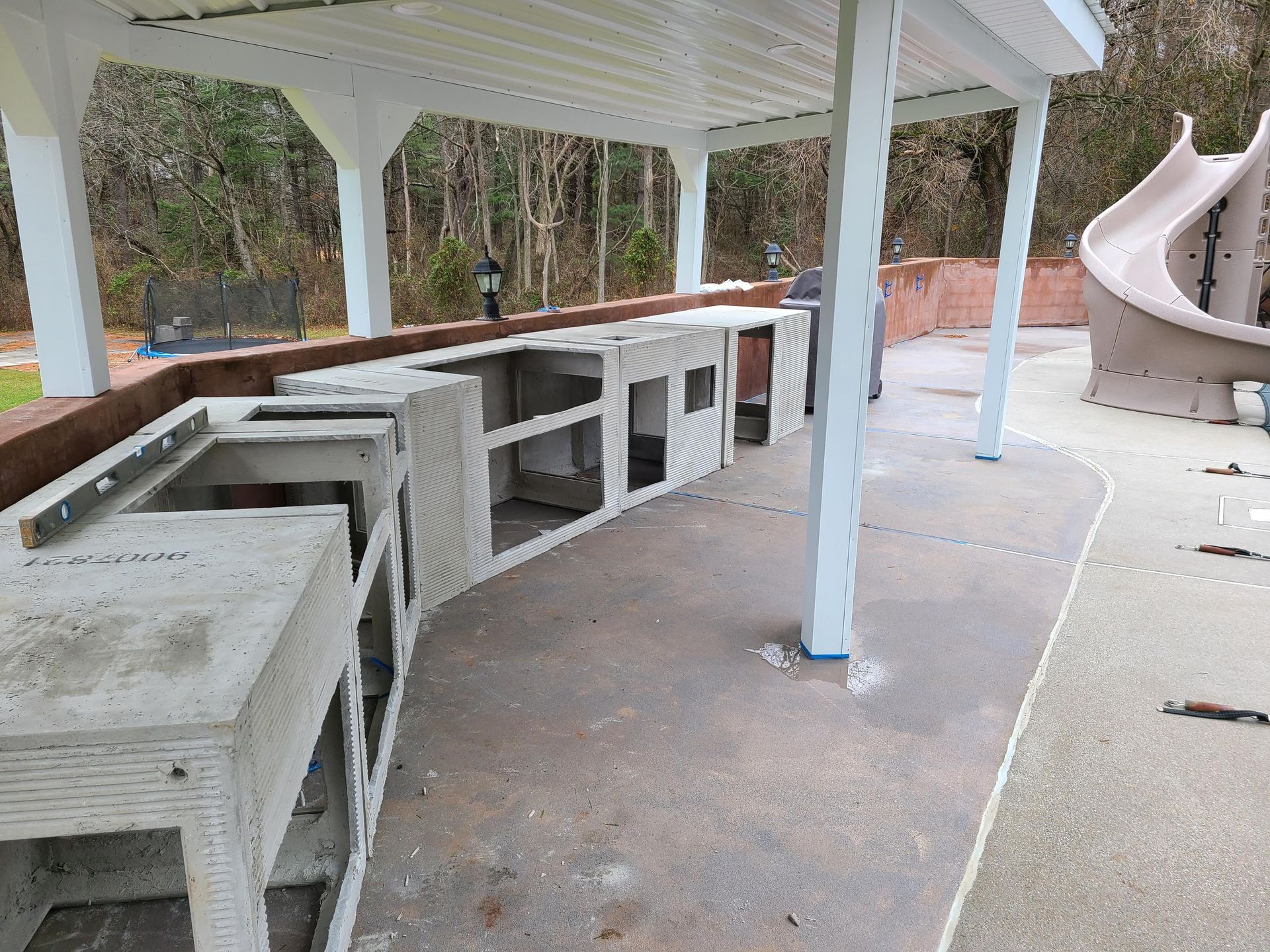 An outdoor concrete kitchen frame under a white pavilion near a curved slide on a patio.