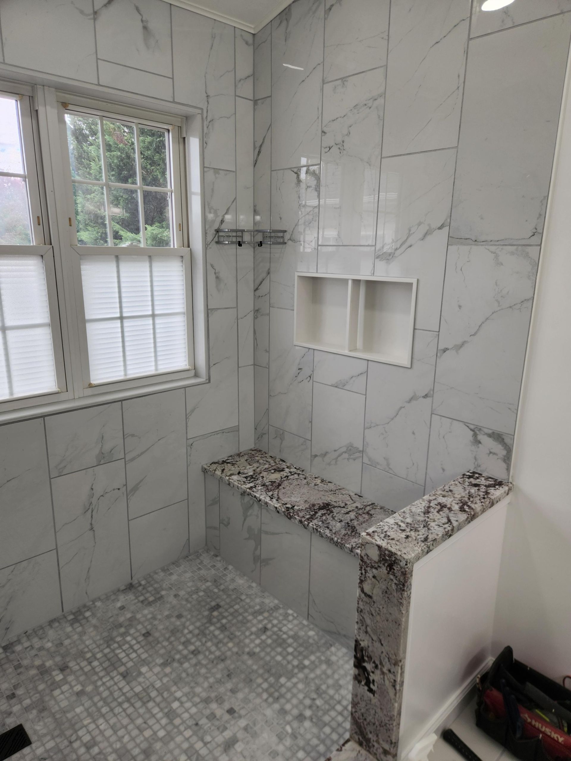 A modern walk-in shower with light grey marble-patterned wall tiles, a mosaic floor, a built-in bench, and a window.
