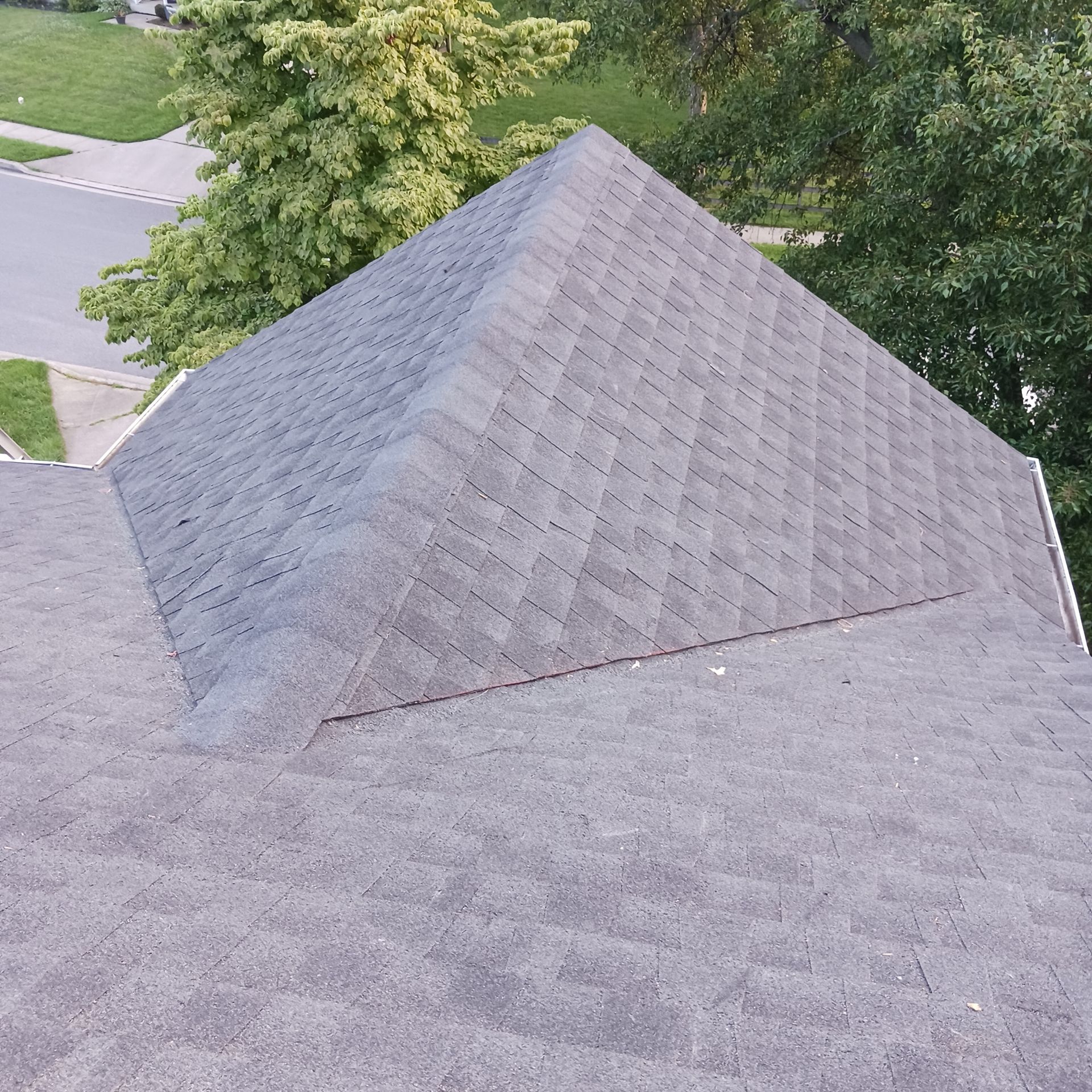 A high-angle view of a gray shingled roof with a prominent hip section, surrounded by trees and a glimpse of a road.