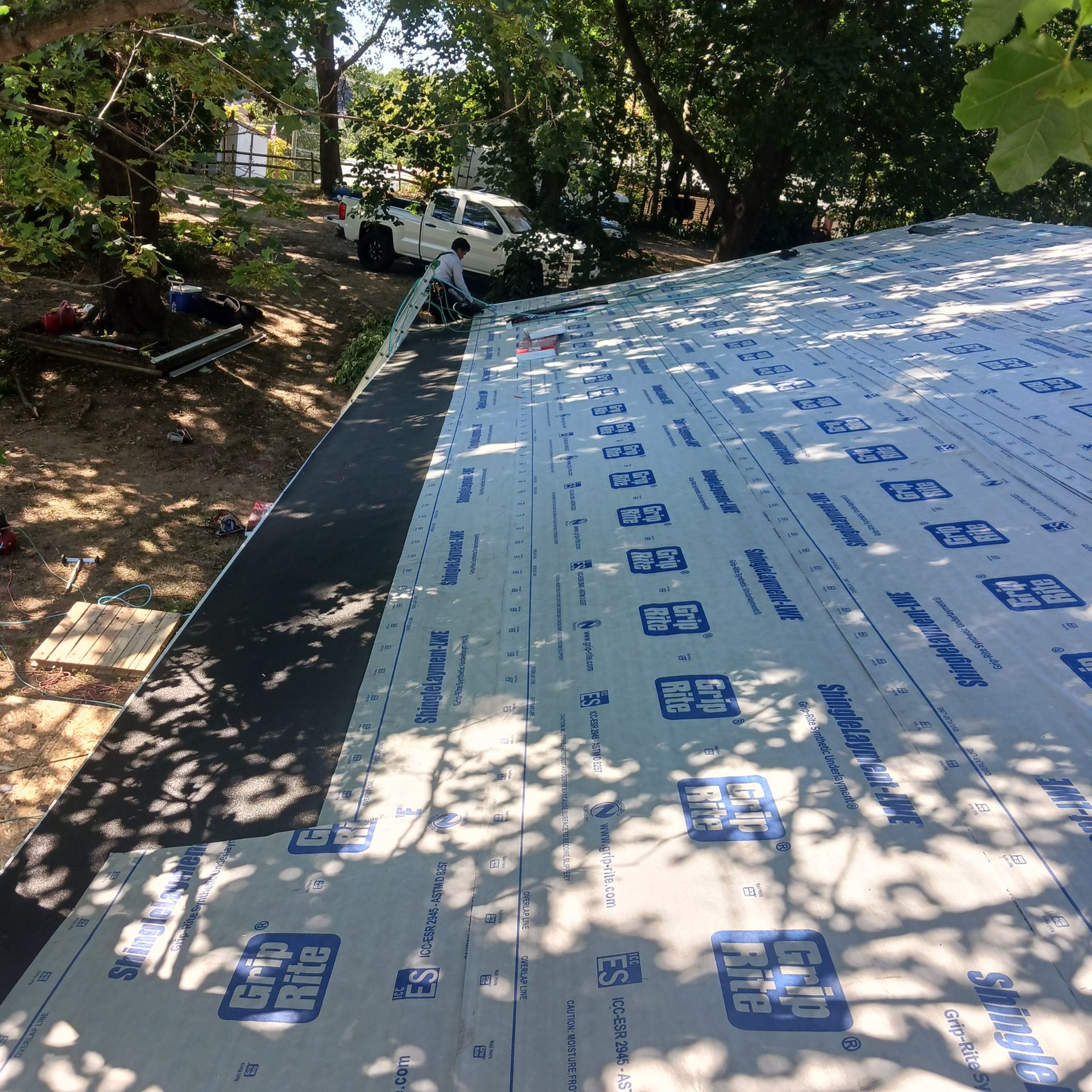A worker installs gray underlayment on a roof, with a white truck parked nearby under trees.
