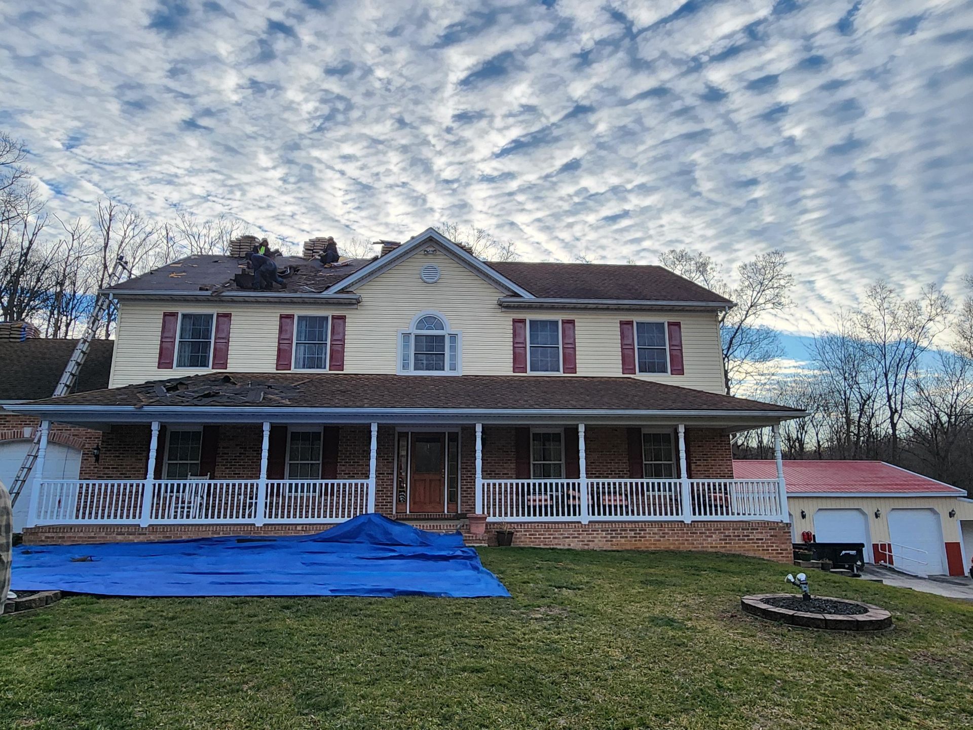 Two-story house with damaged roofing covered by a blue tarp, set under a sky with patterned clouds.