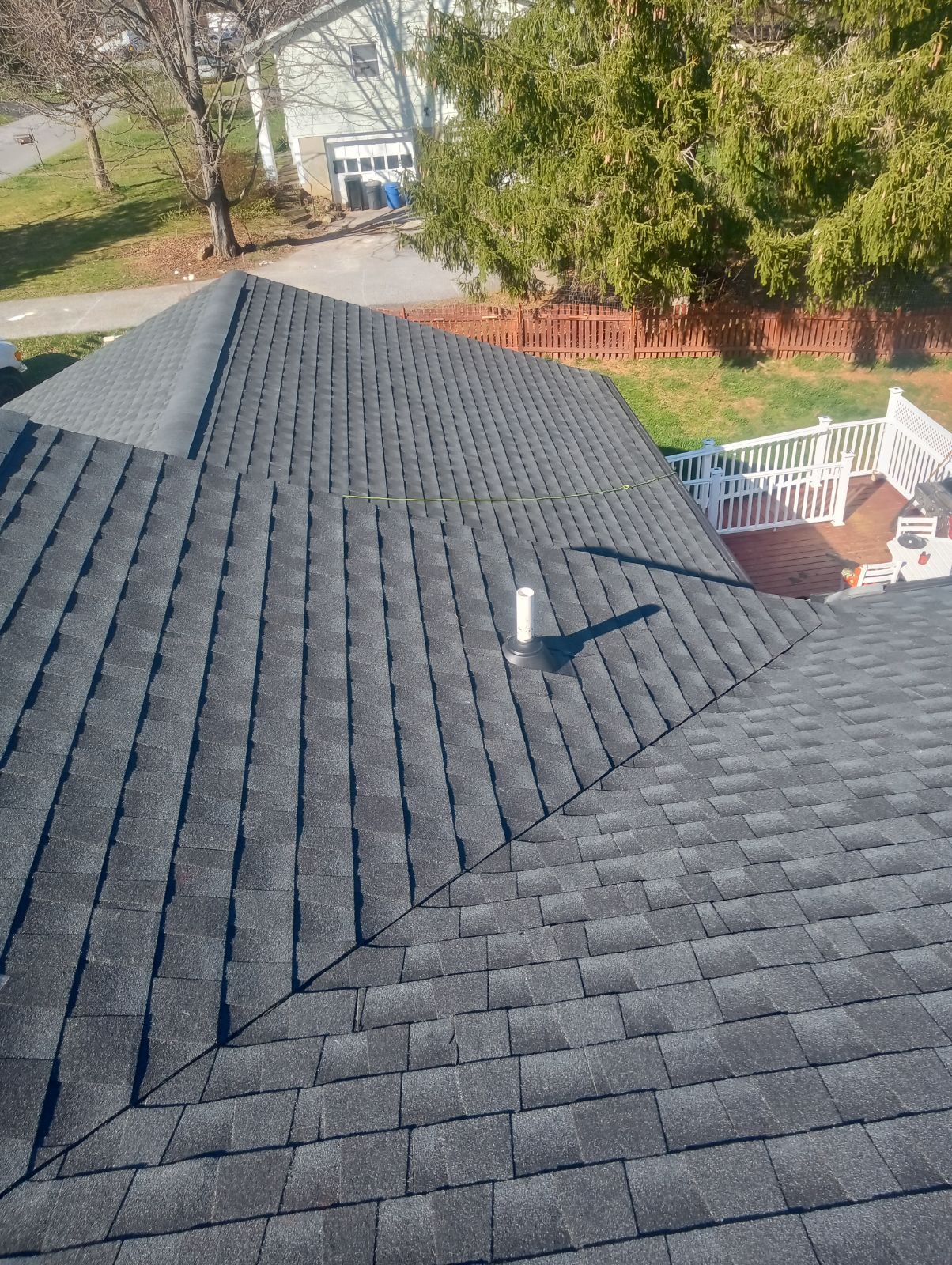 A high-angle view of a grey shingled roof with a vent pipe, looking down onto a backyard, deck, and a neighboring house.