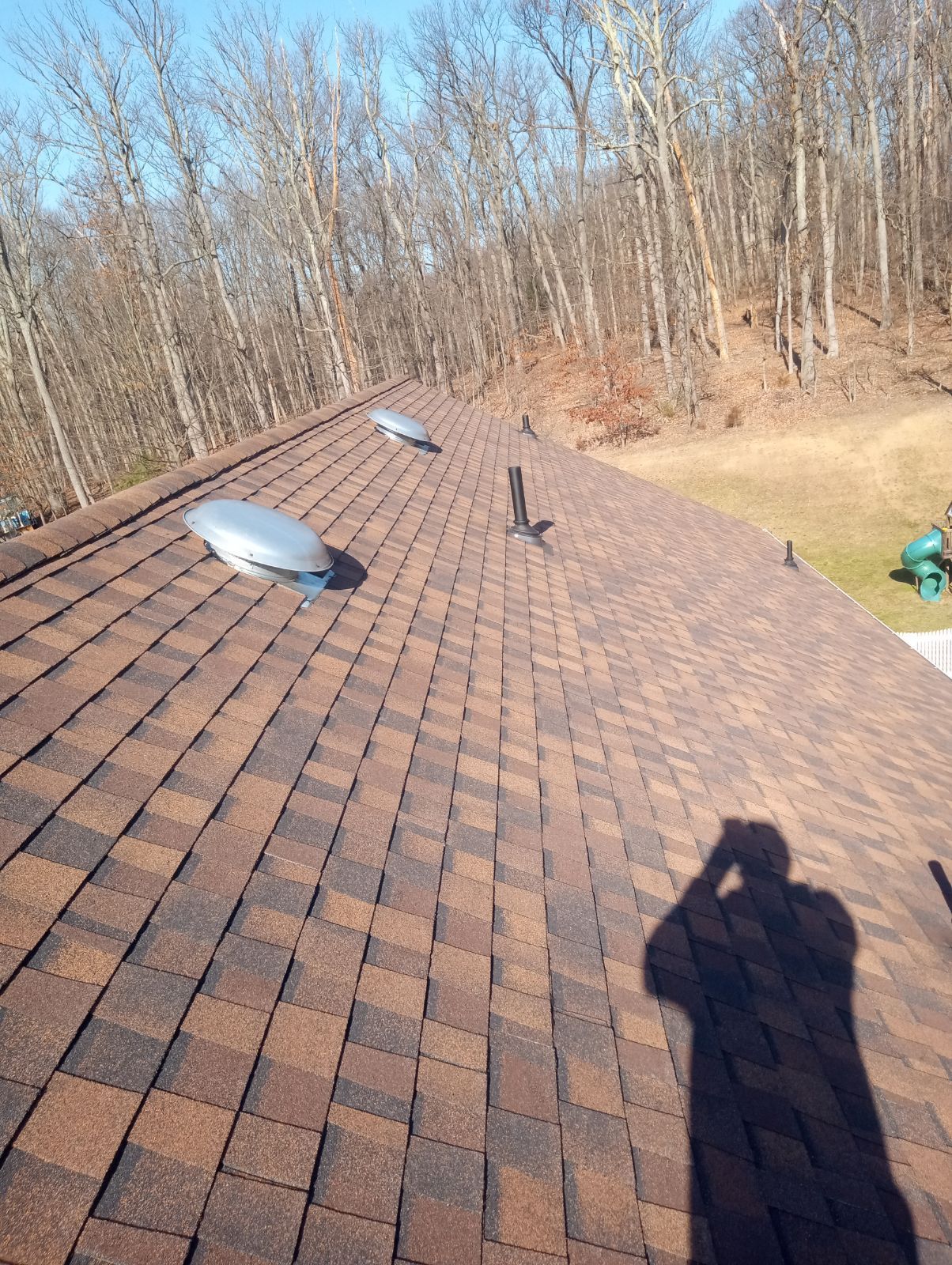 A sunlit view of a brown shingled roof with two metal vents, taken from an elevated position with a shadow in the foreground.