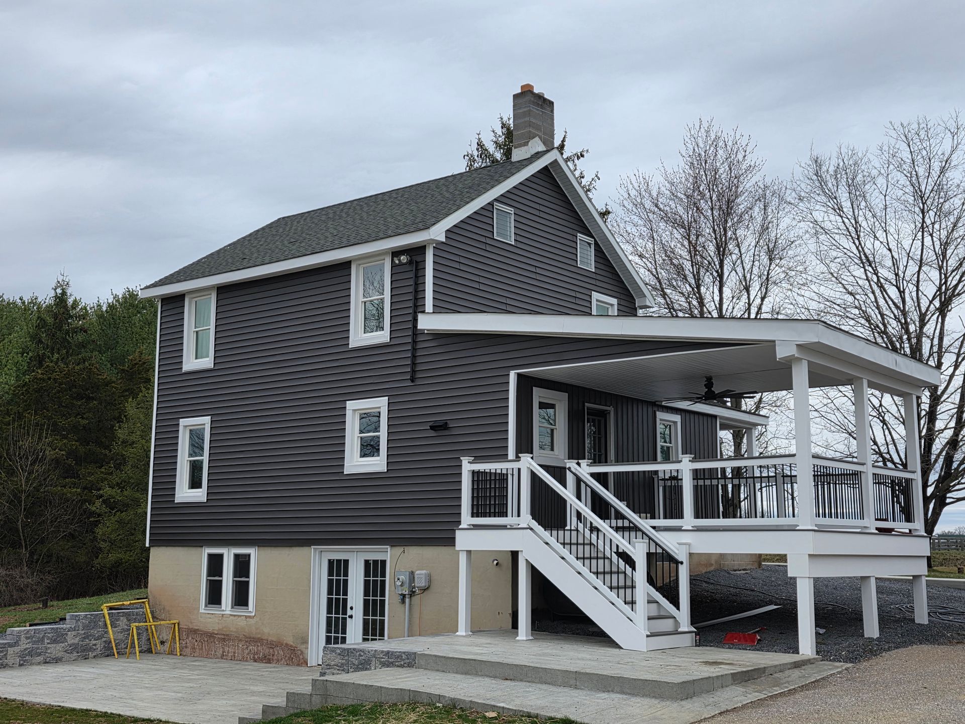 A two-story house with dark gray siding, a stone-colored foundation, and a white deck with stairs leading to a stone patio.