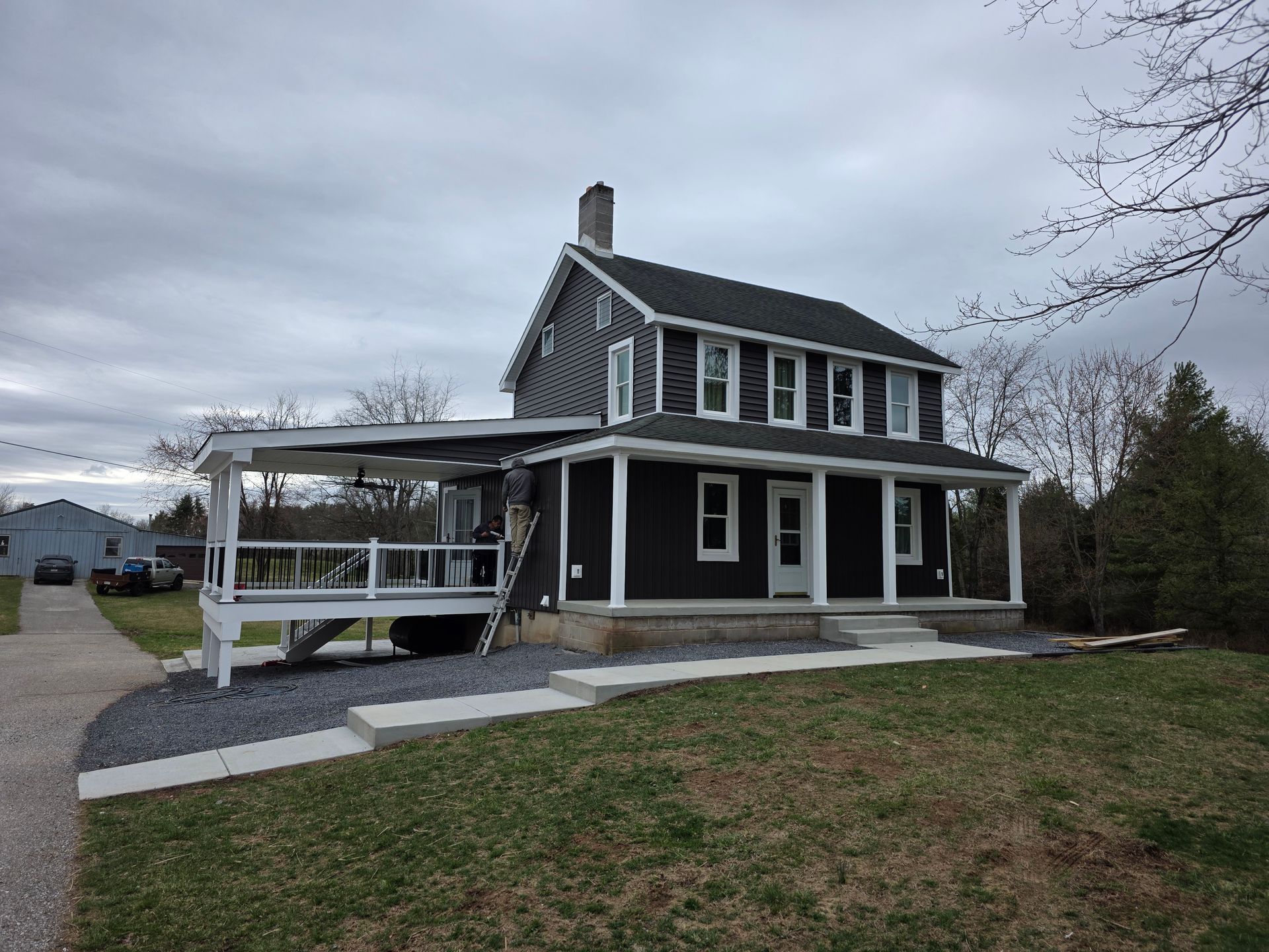 A two-story dark-sided house with a white front porch and a gravel path leading to the entrance under a cloudy sky.