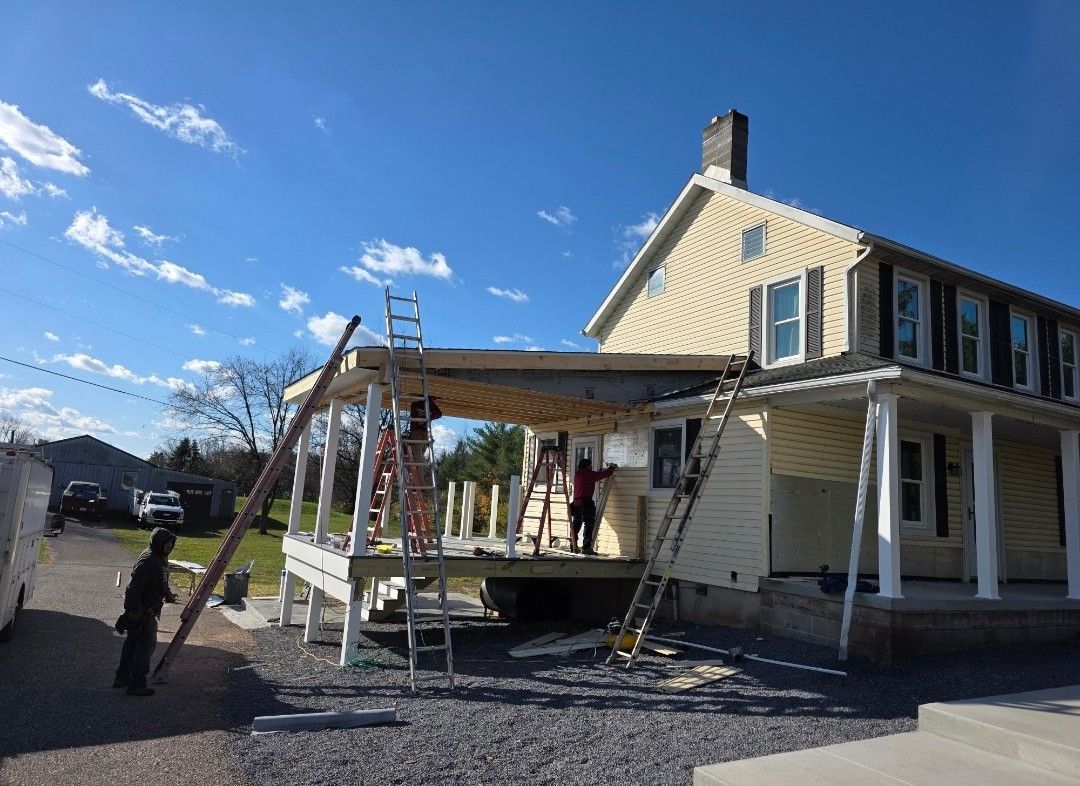 Workers repair a porch on a yellow, two-story house under a bright blue, sunny sky.