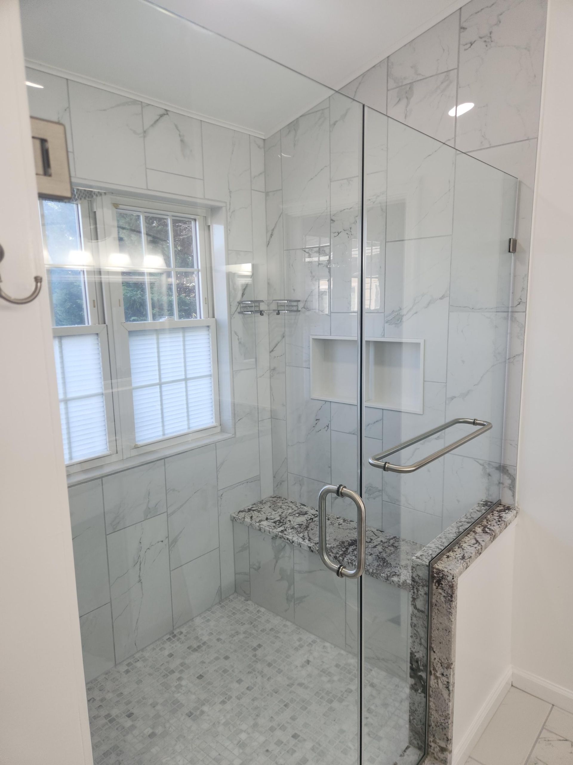 A tiled walk-in shower with a glass door, built-in bench, and a window in a white bathroom.