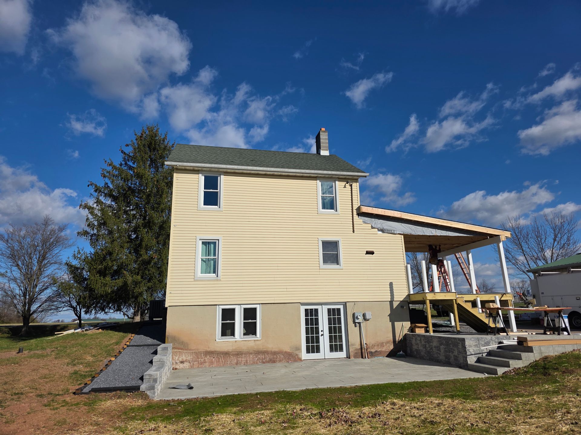 A yellow two-story house with a partially constructed deck and a new stone patio against a blue sky with scattered clouds.