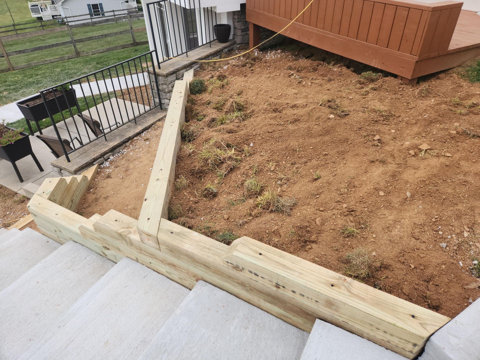 A wooden retaining wall borders a sloped dirt patch next to a concrete staircase and a brown wooden deck.