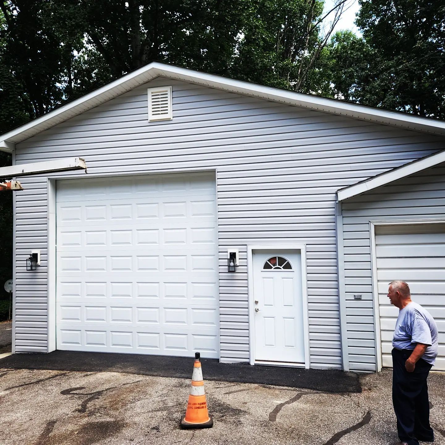 A gray garage with a white door and side entry, a traffic cone in front, and a person standing on the right.
