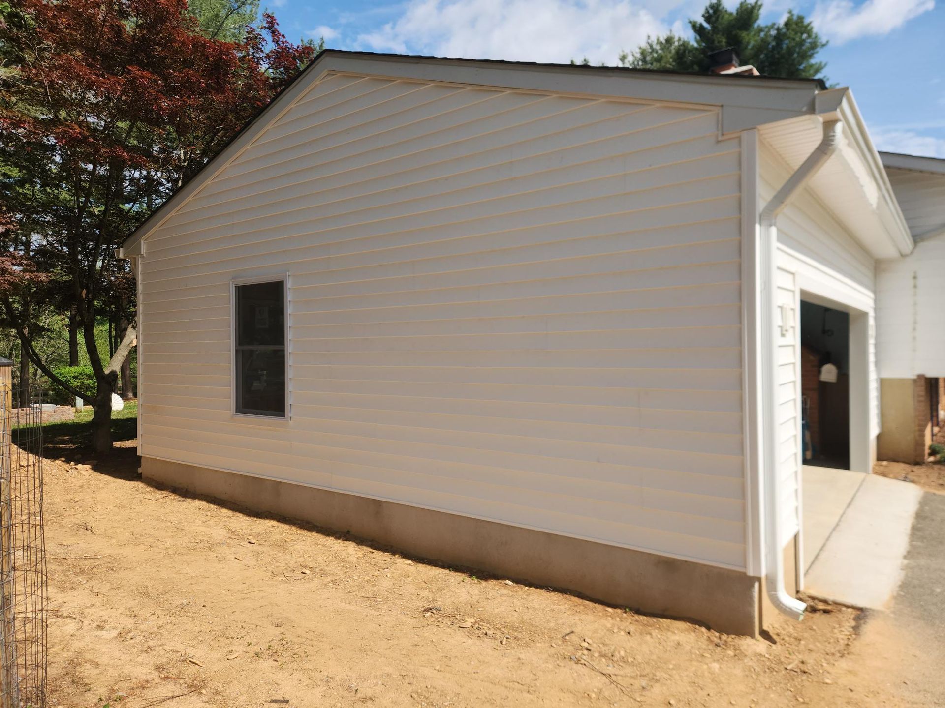 Side view of a garage with light-colored siding, a small window, and a concrete foundation on a dirt lot.