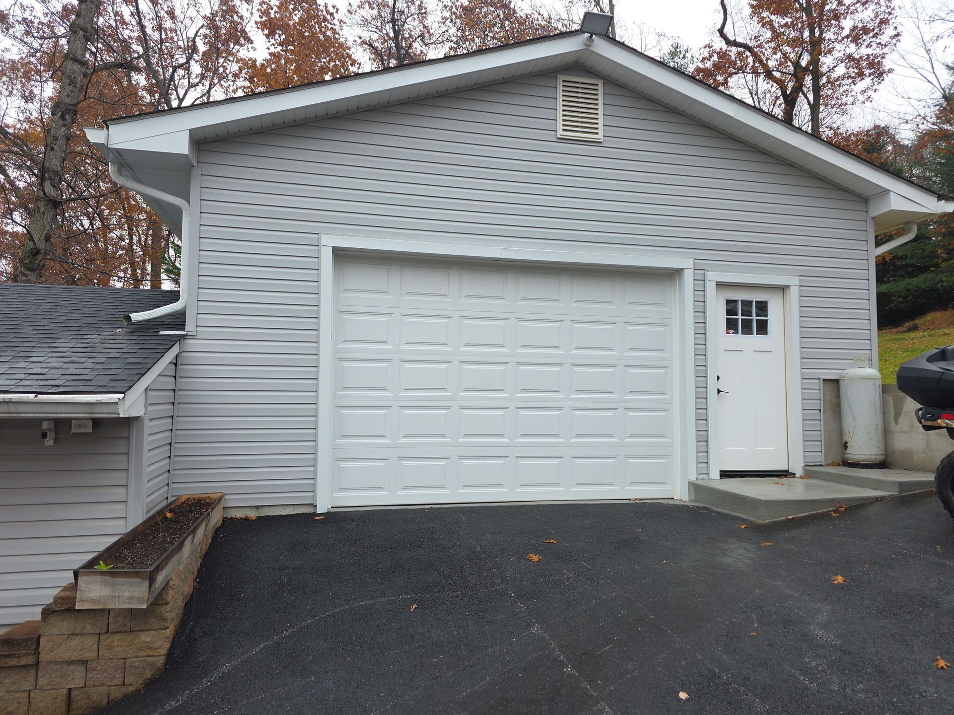 A grey detached garage with a white roll-up door, a side entry door, and a slanted driveway in a wooded setting.
