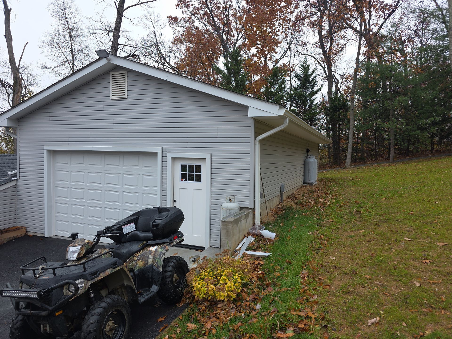 A camouflage ATV parked in front of a white detached garage with a white entry door on a cloudy autumn day.