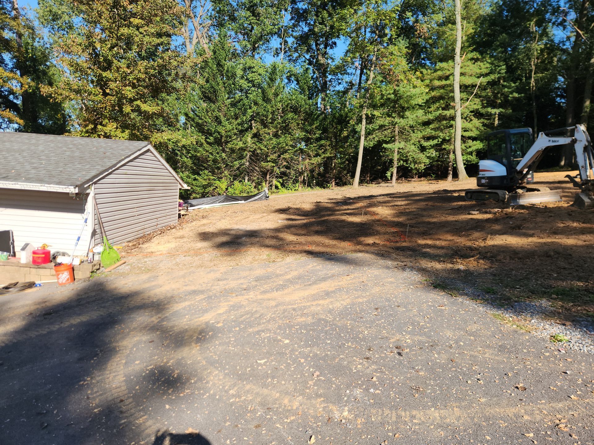 A white mini-excavator sits on a cleared, mulched lot next to a light-colored house at the edge of a wooded area.