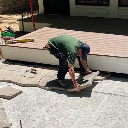 A worker in a green shirt places a large stone paver on a patio in front of a deck.
