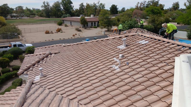 Roofers repairing a tan tile roof, with one worker on the ridge and a suburban street below.
