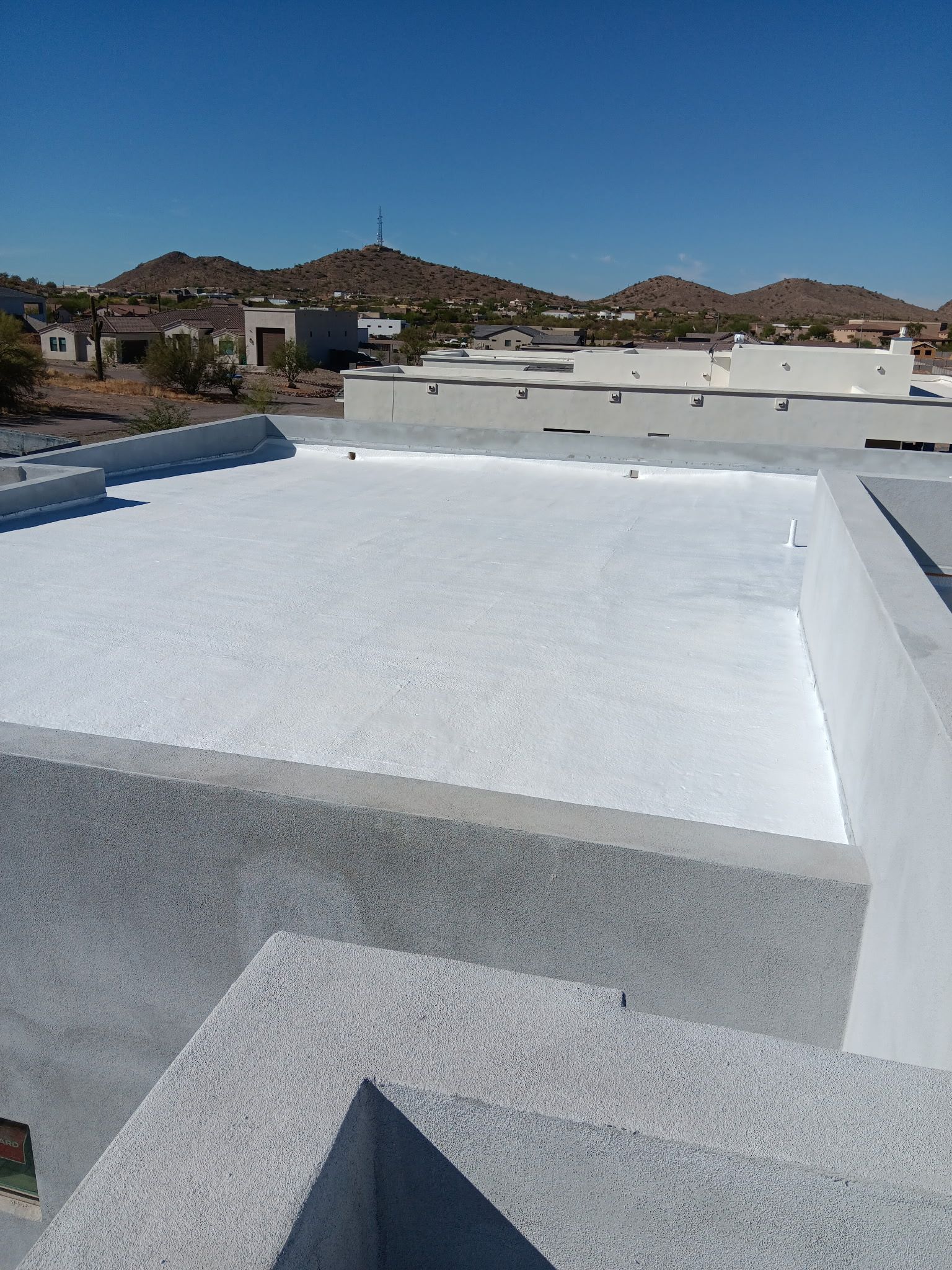 White rooftop terrace with low walls, overlooking desert hills and neighboring buildings under a clear sky
