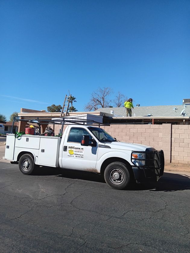 White utility truck parked beside a tan wall, with a worker on the roof under a clear blue sky.