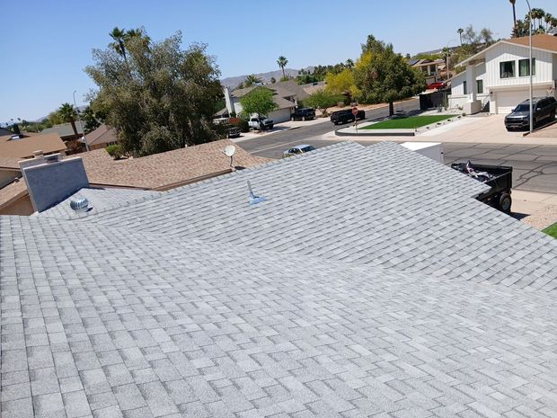 Close-up of a gray shingle roof with houses and trees in a suburban neighborhood under a clear sky