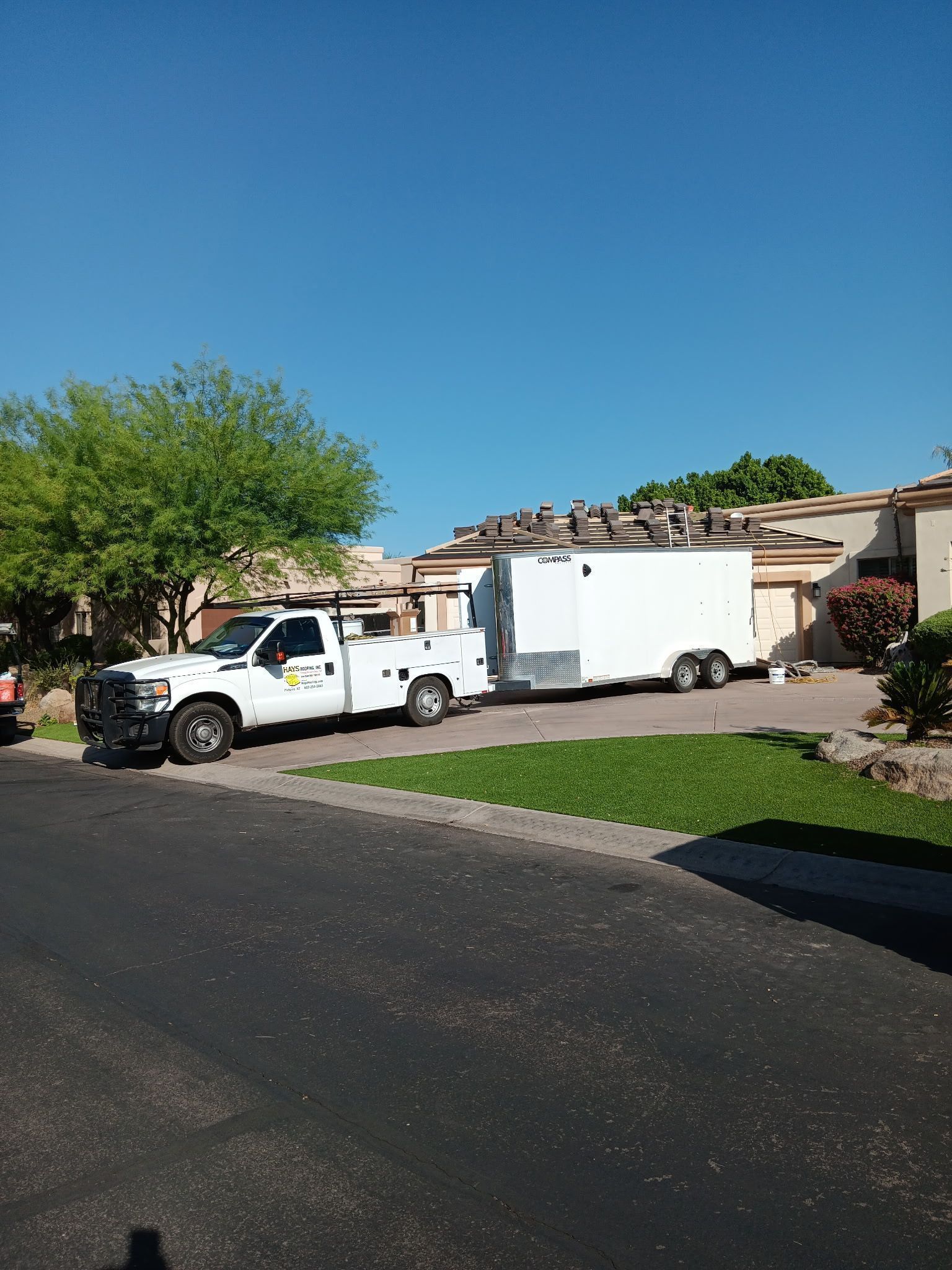 White utility truck and trailer parked on a suburban street beside a driveway under a clear blue sky.