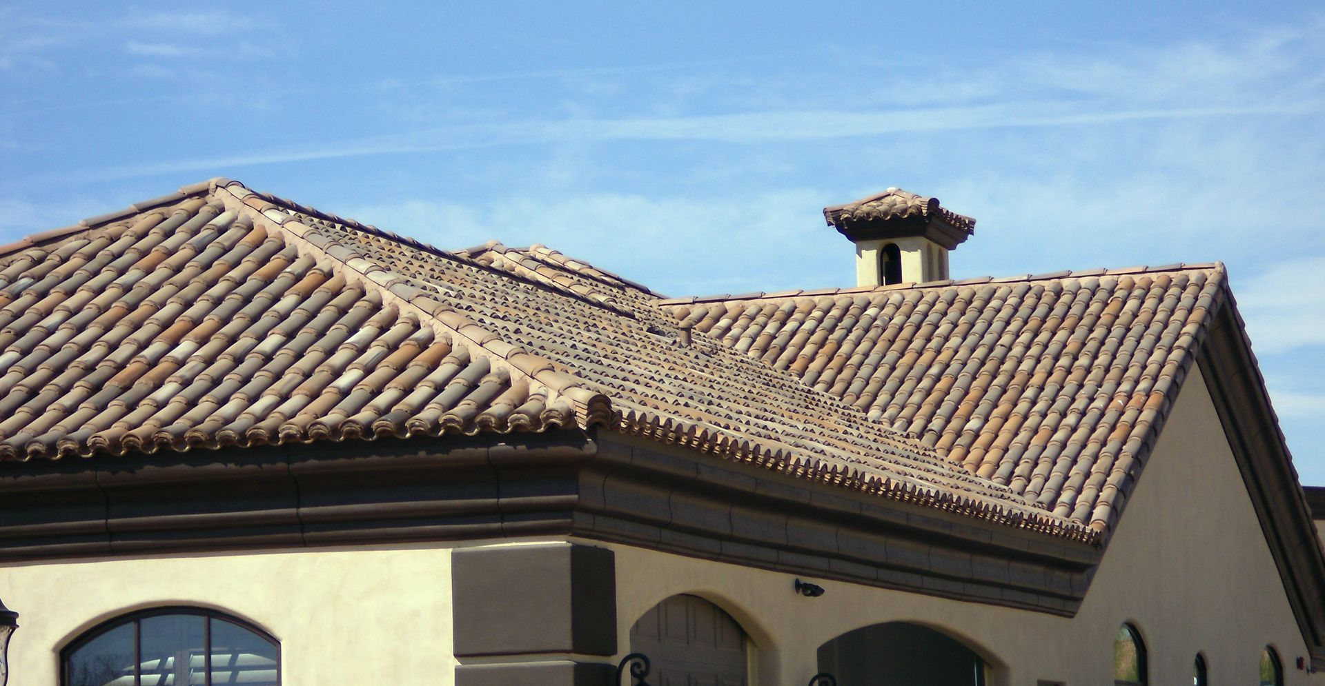 Terracotta tile roof of a stucco house under a blue sky, with a small chimney vent.