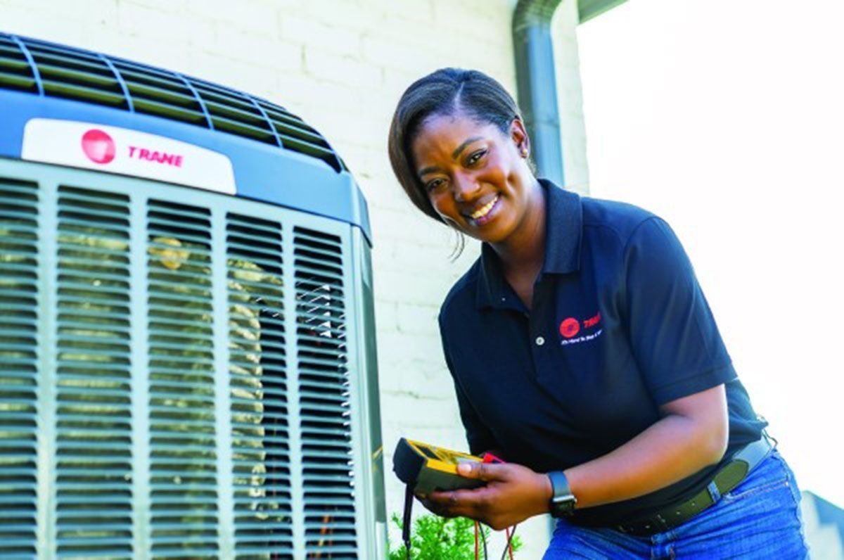 Smiling Black woman in uniform checking an AC unit with a voltmeter outdoors.