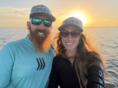 Smiling couple wearing hats and sunglasses on a boat at sunset, with ocean behind them