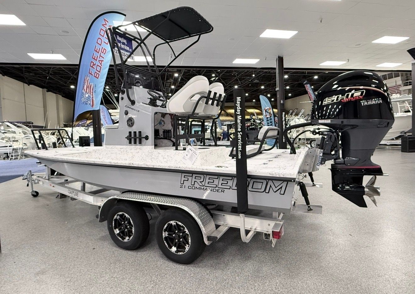 White fishing boat on a trailer indoors at a boat show, with a black motor and canopy.