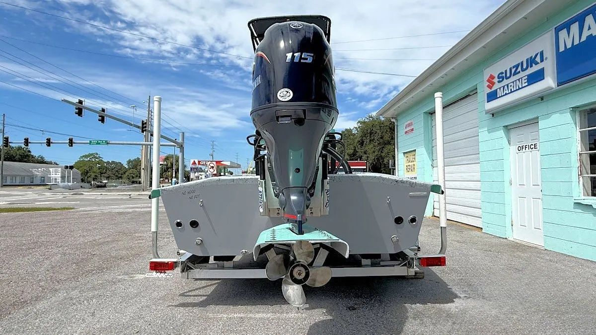 Large black boat motor on a trailer in a gravel lot beside a turquoise building under a blue sky.