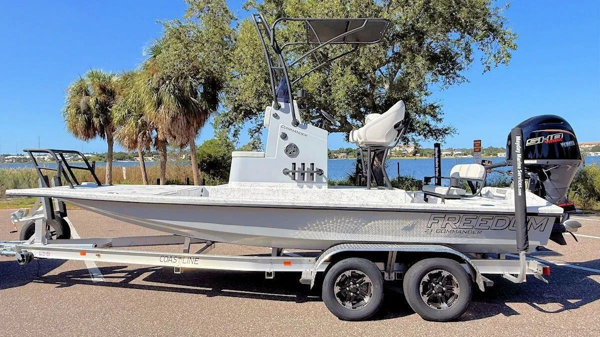 White center-console fishing boat on a trailer parked outdoors near palm trees and water.