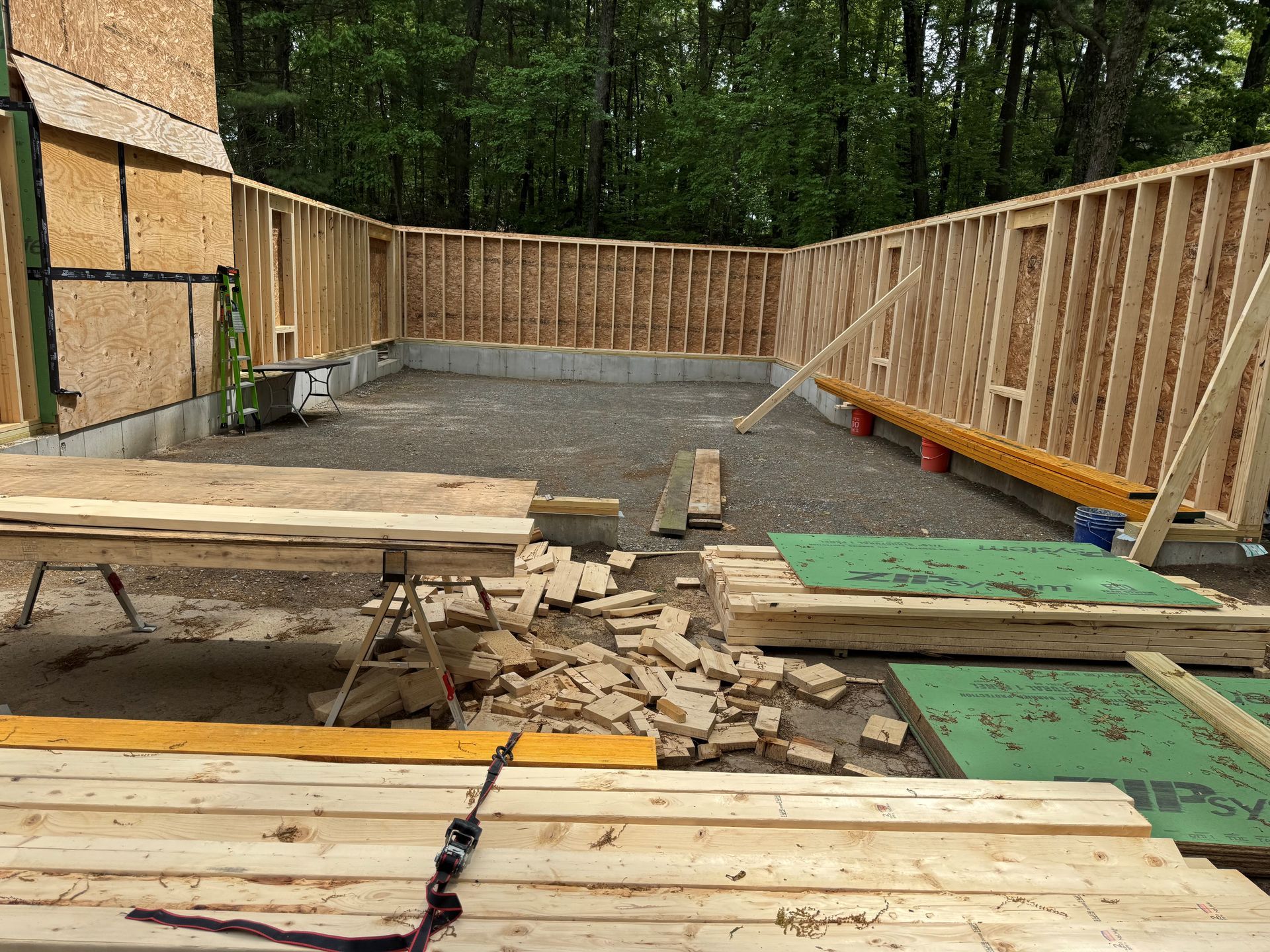 Framed wooden house under construction with scattered lumber and green floor panels in a wooded area