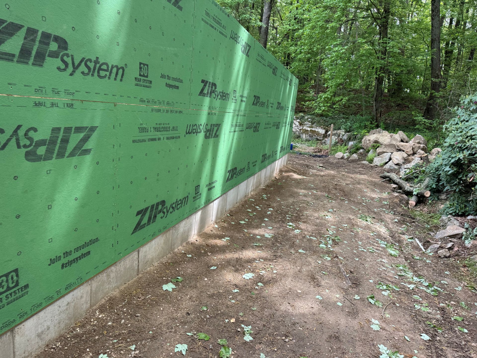A wooded path beside a green Zip System wall with exposed soil and rocks.