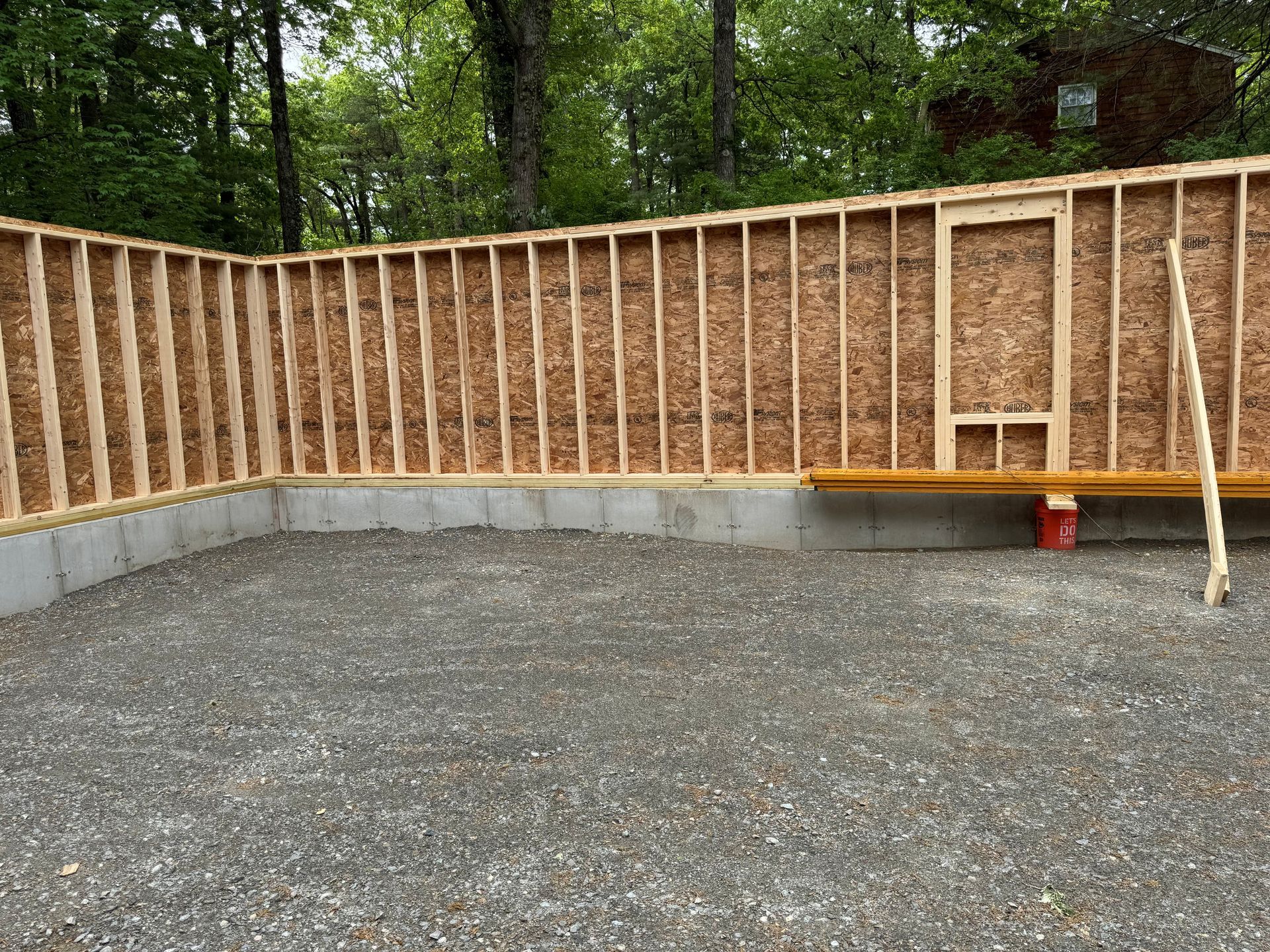 Framed wooden shed walls on a gravel base, with a concrete foundation and trees behind the site