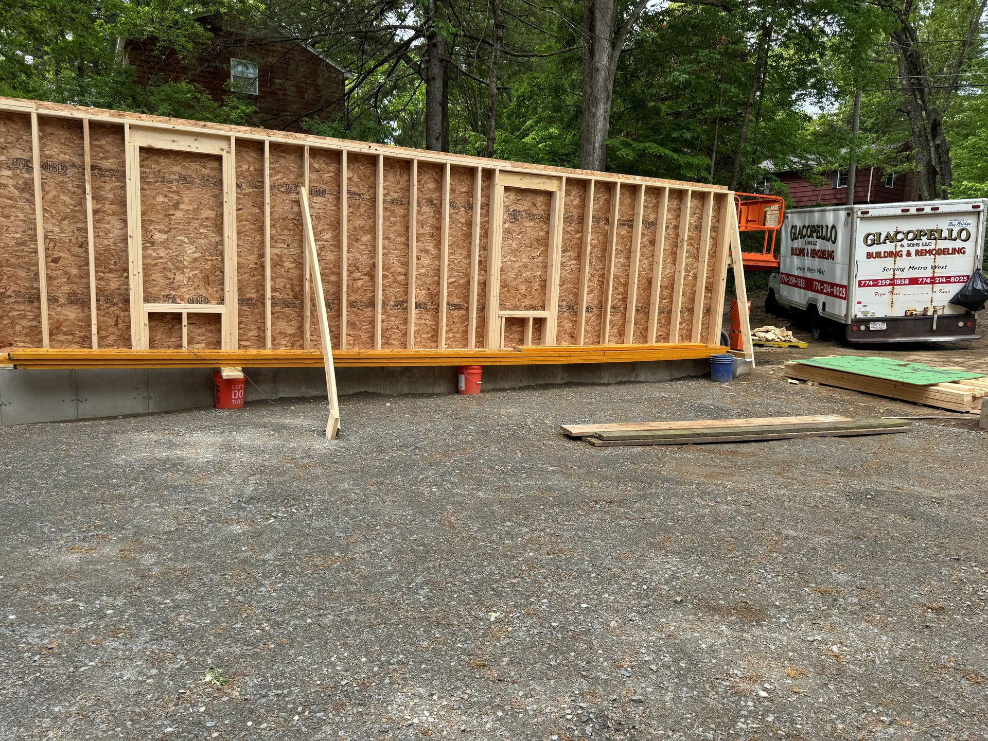 Large wooden wall frame on gravel beside a tree-lined worksite, with a truck and trailer in the background