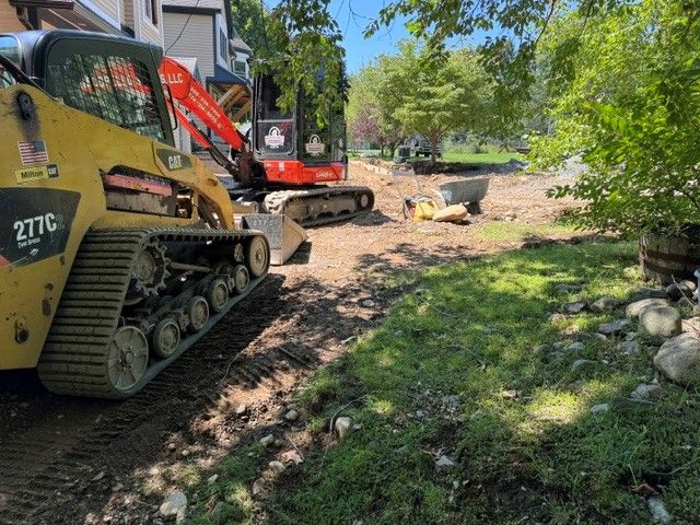 Two excavators on a muddy dirt path at a sunny construction site, with trees and grass nearby