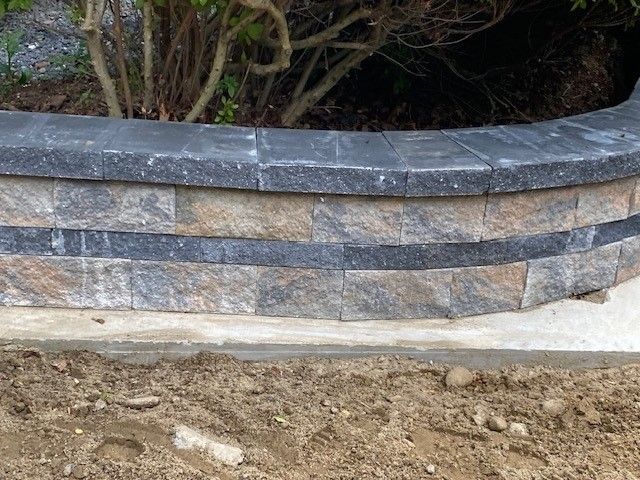 Curved stone retaining wall with gray and tan blocks beside a dirt path and shrubs