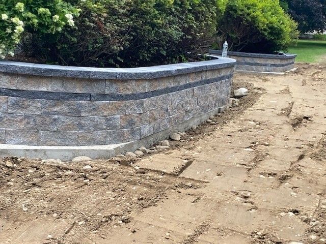 Stone retaining wall along a dirt path beside dense green shrubs and trees