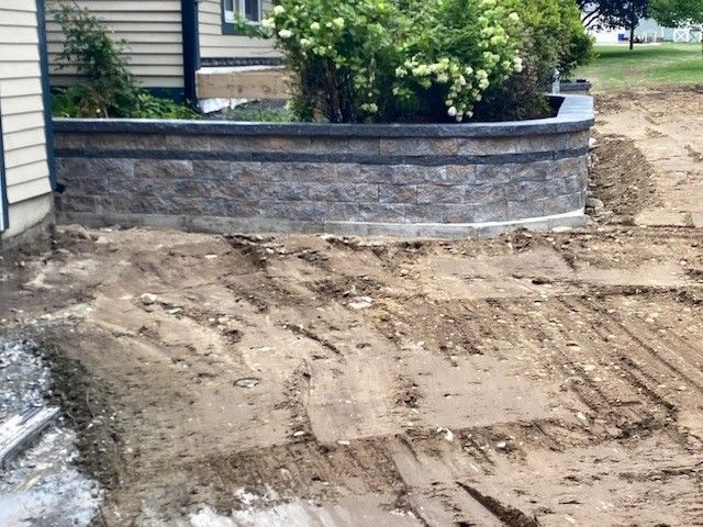 Dirt-filled driveway under construction beside a gray stone retaining wall and shrubs