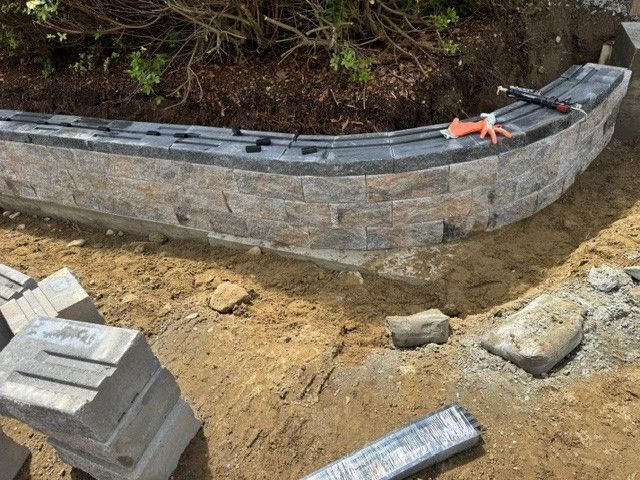 Curved stone retaining wall under construction along a dirt slope, with blocks and tools nearby