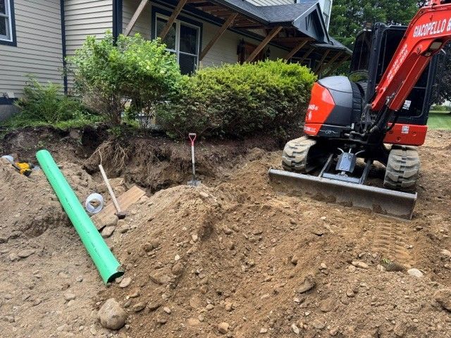 Excavator beside a house digging a dirt trench with green and white pipes nearby