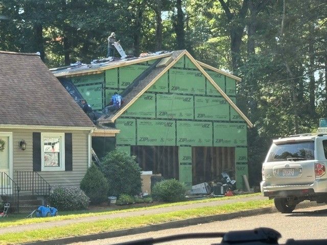 House under construction with green siding wrap, a ladder on the roof, and a white SUV parked nearby.